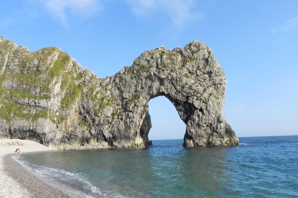 The limestone arch of Durdle door, with grey almost vertical rocks, with some mossy grassy vegetation on the top, with a backdrop of a cloudless blue sky, pebble beach and blue sea. 